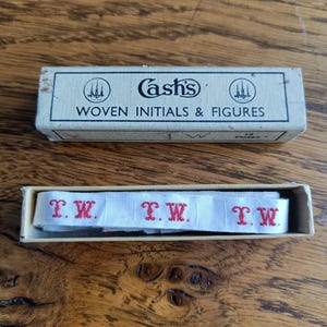 Pode incluir: Uma caixa vintage de iniciais e figuras tecidas da Cash's. A caixa contém uma fita branca com as iniciais "T.W." em vermelho. A caixa é bege com texto preto que diz "Cash's Woven Initials & Figures."