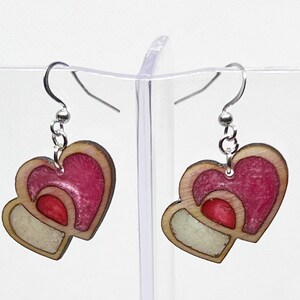 May include: A pair of dangle earrings featuring two layered heart shapes made of wood. The hearts are painted in shades of pink and white.