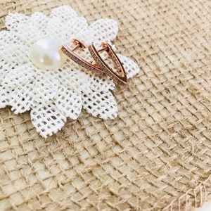 May include: Rose gold earrings with a V-shaped design and clear crystal accents. The earrings are on a white fabric background.
