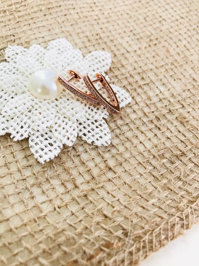 May include: Rose gold earrings with a V-shaped design and clear crystal accents. The earrings are on a white fabric background.