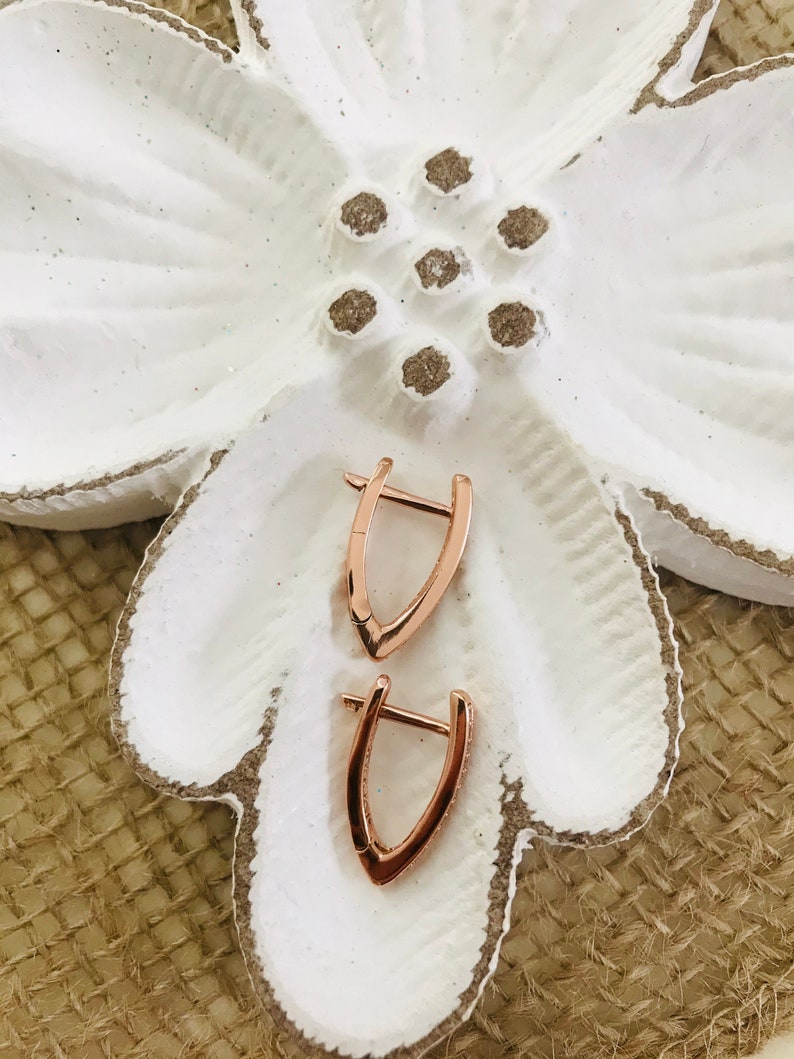 May include: A pair of rose gold hoop earrings with a v-shaped design. The earrings are on a white flower-shaped dish.