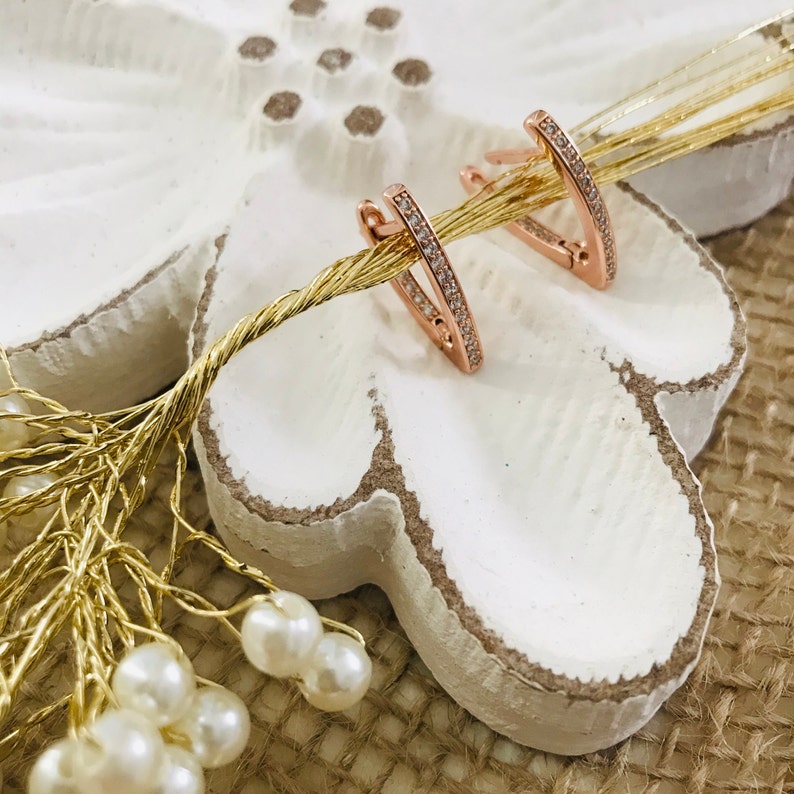 May include: A pair of rose gold hoop earrings with a row of clear cubic zirconia stones around the edge.