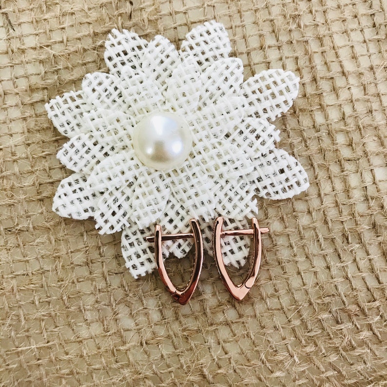 May include: A pair of rose gold hoop earrings with a delicate, curved design. The earrings are displayed on a burlap background with a white fabric flower.