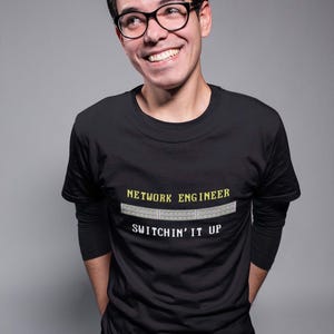 Puede incluir: Un hombre con una camiseta negra de manga larga con el texto "NETWORK ENGINEER" y "SWITCHIN' IT UP" en una fuente pixelada. El texto está acompañado por un gráfico de un conmutador de red.