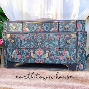 May include: A black chest of drawers with four drawers features a blue and pink floral pattern with birds. The dresser has gold-coloured knobs and a curved front.