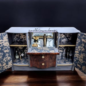 May include: A grey bar cabinet with two doors and a mirrored back. The doors are open to reveal shelves lined with gold and blue patterned paper. The cabinet has a wooden top with two drawers and gold accents.