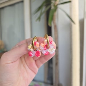 Könnte beinhalten: Goldene Creolen-Ohrringe, verziert mit Clustern aus durchscheinenden, blütenförmigen Anhängern. Die Anhänger weisen Farbtöne von Rosa, Orange und Goldblatt-Akzente auf. Diese Ohrringe bieten einen zarten und femininen Touch.