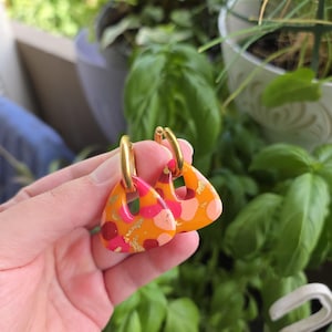 Könnte beinhalten: Ein Paar Creolen mit dreieckigen orangefarbenen Anhängern, die ein buntes Muster aus rosa, roten und hellorangen Punkten aufweisen und mit kleinen Goldfolienakzenten versehen sind. Die Anhänger hängen an polierten goldfarbenen Metallringen.