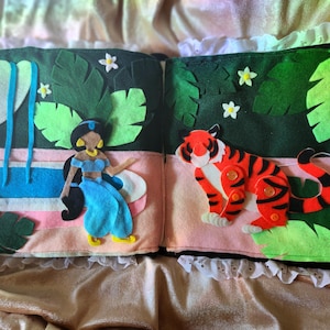 Puede incluir: Un libro de fieltro con dos páginas. La página izquierda presenta un recorte de fieltro de la princesa Jasmine de Aladdin, con un atuendo azul y un collar de oro. Ella está de pie frente a una piscina azul con una cascada. La página derecha presenta un recorte de fieltro de un tigre con rayas naranjas y negras. El tigre está de pie sobre una hoja verde.