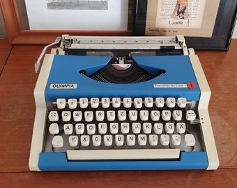May include: A blue and white Olympia Traveller de Luxe typewriter with a black keyboard and a red number 1 on the side. The typewriter has a white carriage return lever and a black roller.