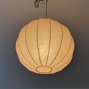 Könnte beinhalten: Eine runde, cremefarbene Pendelleuchte mit geripptem Design. Das Licht hängt an einer Metallhalterung, die an der Decke befestigt ist. Das Licht ist eingeschaltet und beleuchtet den Raum.