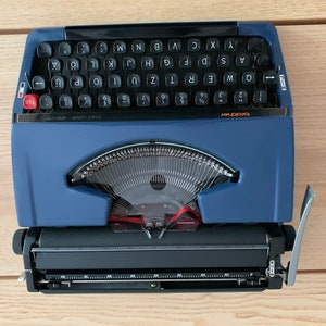 Puede incluir: Una m&aacute;quina de escribir vintage azul y negra con una tecla de tabulaci&oacute;n roja y el texto "brother deluxe 501" en la parte delantera.