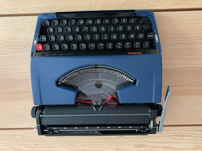 Puede incluir: Una m&aacute;quina de escribir vintage azul y negra con una tecla de tabulaci&oacute;n roja y el texto "brother deluxe 501" en la parte delantera.