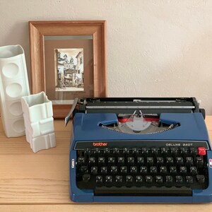 Puede incluir: Una m&aacute;quina de escribir vintage azul con un teclado negro y el texto "brother DELUXE 240T" en la parte delantera. La m&aacute;quina de escribir est&aacute; sobre una superficie de madera con dos jarrones de cer&aacute;mica blanca.