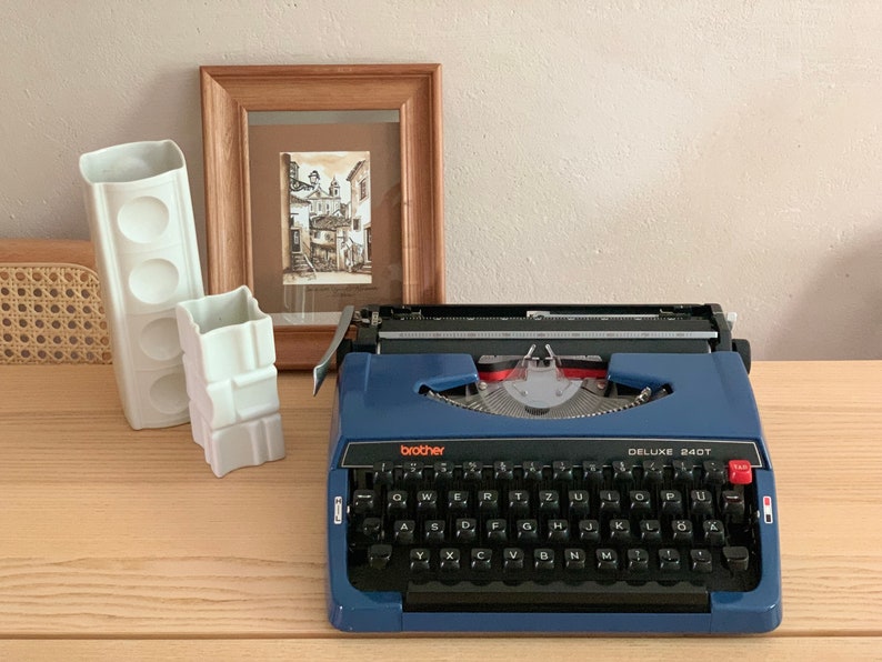 Puede incluir: Una m&aacute;quina de escribir vintage azul con un teclado negro y el texto "brother DELUXE 240T" en la parte delantera. La m&aacute;quina de escribir est&aacute; sobre una superficie de madera con dos jarrones de cer&aacute;mica blanca.