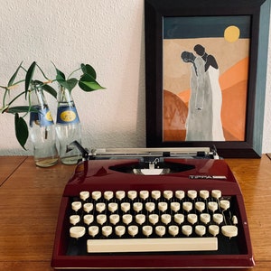 May include: A vintage red typewriter with a cream-coloured keyboard and the word "TIPPA" printed on the front. The typewriter is sitting on a wooden table next to two clear glass bottles and a framed print of two figures in a desert landscape.