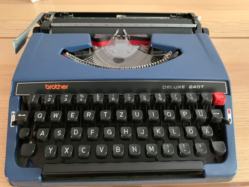 Puede incluir: Una m&aacute;quina de escribir Brother Deluxe 240T azul con un teclado negro y una tecla de tabulaci&oacute;n roja.