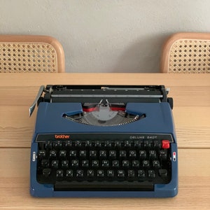 Puede incluir: Una m&aacute;quina de escribir Brother Deluxe 240T azul con un teclado negro y una tecla de tabulaci&oacute;n roja. La m&aacute;quina de escribir tiene una palanca de retorno de carro plateada y una palanca de liberaci&oacute;n de papel plateada.