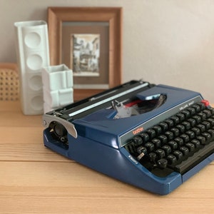 Puede incluir: Una m&aacute;quina de escribir Brother Deluxe 1407 azul con un teclado negro y acentos rojos. La m&aacute;quina de escribir est&aacute; sobre una superficie de madera.