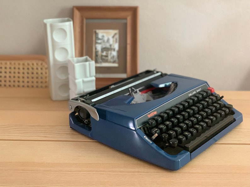 Puede incluir: Una m&aacute;quina de escribir Brother Deluxe 1407 azul con un teclado negro y acentos rojos. La m&aacute;quina de escribir est&aacute; sobre una superficie de madera.