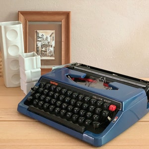 Puede incluir: Una m&aacute;quina de escribir Brother Deluxe 240T azul con un teclado negro y letras naranjas. La m&aacute;quina de escribir est&aacute; sobre una superficie de madera.