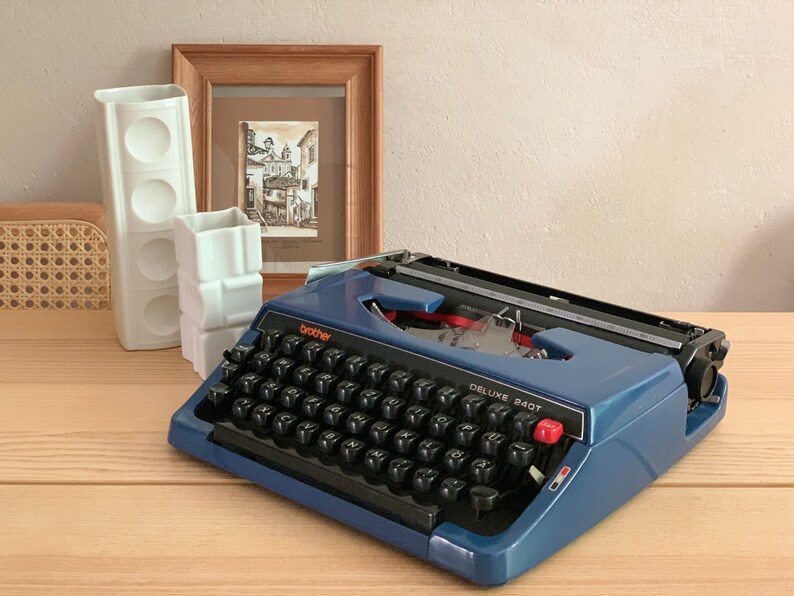 Puede incluir: Una m&aacute;quina de escribir Brother Deluxe 240T azul con un teclado negro y letras naranjas. La m&aacute;quina de escribir est&aacute; sobre una superficie de madera.
