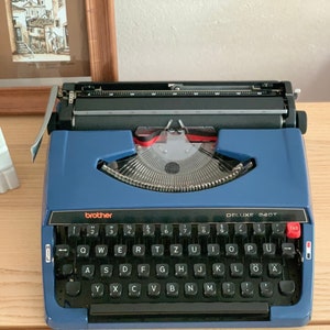 May include: A blue Brother Deluxe 240T typewriter with a black keyboard and red accents. The typewriter has a tab key and a carriage return lever.