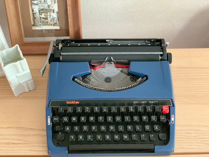 Puede incluir: Una m&aacute;quina de escribir Brother Deluxe 240T azul con un teclado negro y acentos rojos. La m&aacute;quina de escribir tiene una tecla de tabulaci&oacute;n y una palanca de retorno de carro.