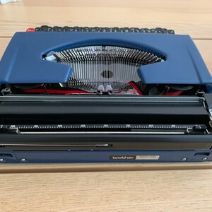 Puede incluir: Una m&aacute;quina de escribir Brother azul con un carro negro y una palanca de retorno plateada. La m&aacute;quina de escribir est&aacute; abierta y lista para escribir.