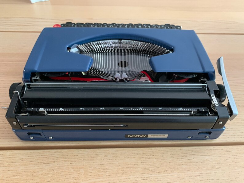 Puede incluir: Una m&aacute;quina de escribir Brother azul con un carro negro y una palanca de retorno plateada. La m&aacute;quina de escribir est&aacute; abierta y lista para escribir.