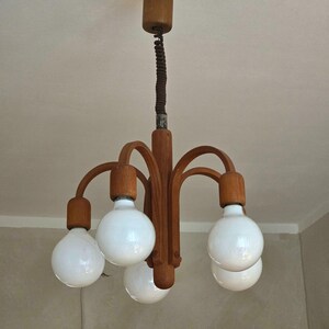 Puede incluir: Lámpara de araña de madera con cinco bombillas globulares blancas. La lámpara tiene una columna central de madera con brazos curvos que sostienen las bombillas. La lámpara está suspendida del techo por un cable marrón.
