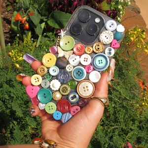 May include: A clear phone case adorned with a variety of colorful buttons in shades of blue, green, red, yellow, and white. The buttons are of different sizes and shapes, creating a unique and textured design. The case is held in front of a lush green background.