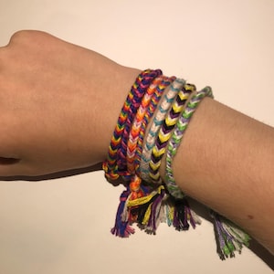 May include: A stack of colourful friendship bracelets with tassels. The bracelets are made of woven string in various colours, including purple, blue, green, yellow, orange, and white.