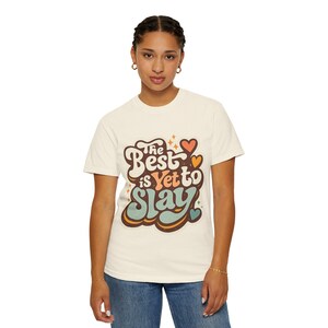 Op de afbeelding: Crèmekleurig T-shirt met de tekst "The Best is Yet to Slay" in een retro lettertype. Het ontwerp bevat oranje, bruine en turquoise accenten, met hart- en stervormen. Het model draagt een blauwe jeans.