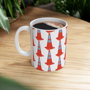 May include: White ceramic coffee mug with a repeating pattern of orange and gray traffic cones. The mug is filled with dark coffee and has a white handle. The mug is sitting on a wooden surface.