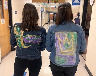 May include: Two denim jackets with hand-painted designs. One features a Mardi Gras mask in green, purple, and gold. The other displays a crown in gold, purple, and green. Both jackets are worn by individuals standing in a hallway.