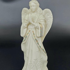 Op de afbeelding: Ivoor engelbeeldje met gevouwen handen, gekleed in een lange jurk met decoratieve patronen. De mouwen en zoom van de jurk van de engel zijn versierd met gouden, groene en paarse accenten. De engel heeft grote, gedetailleerde vleugels.