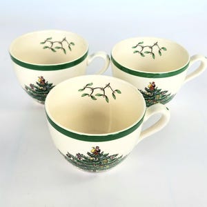 May include: Three white ceramic teacups with green trim and a Christmas tree design. The cups have a traditional Christmas tree design with green leaves, red and yellow ornaments, and a brown bird on top.