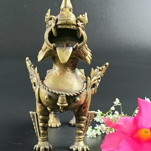 Puede incluir: Una estatua de metal dorado de una criatura m&iacute;tica con una expresi&oacute;n feroz. La criatura tiene una corona en la cabeza y una campana colgando de su cuello. La estatua est&aacute; de pie sobre una superficie negra.