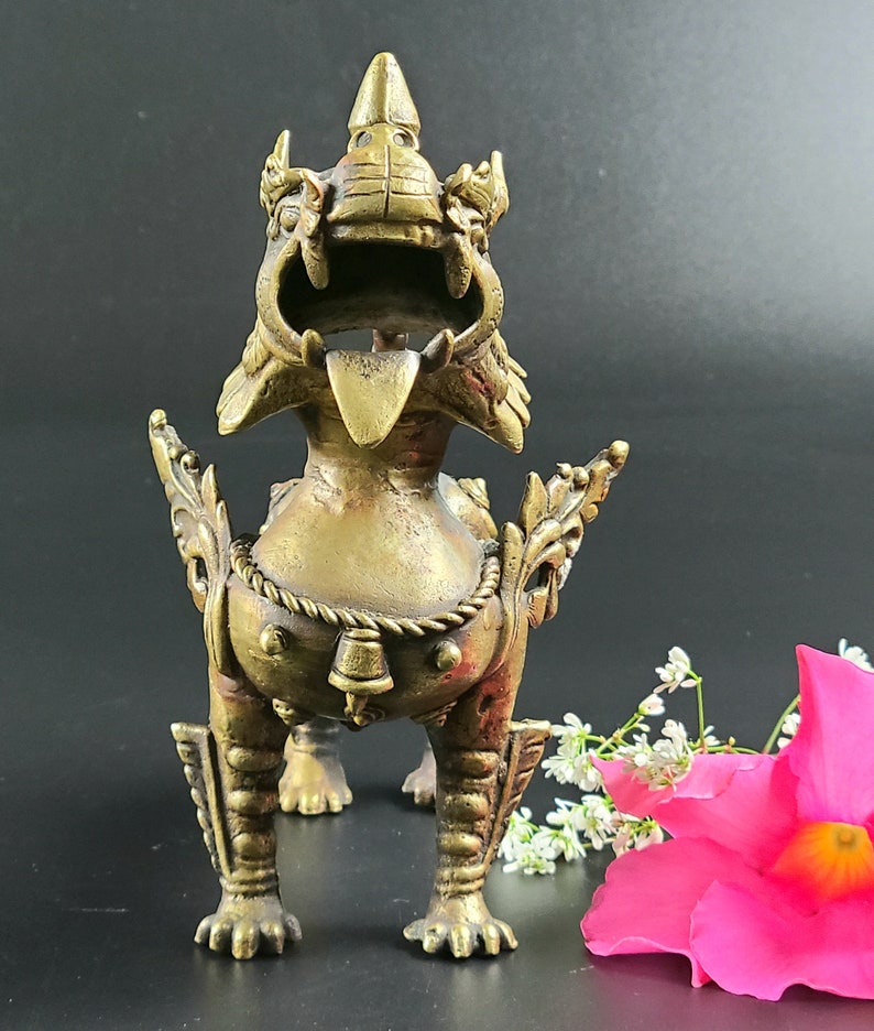 Puede incluir: Una estatua de metal dorado de una criatura m&iacute;tica con una expresi&oacute;n feroz. La criatura tiene una corona en la cabeza y una campana colgando de su cuello. La estatua est&aacute; de pie sobre una superficie negra.