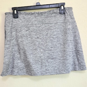 May include: A gray athletic skort hanging on a black hanger. The skort has a built-in shorts layer and a flared skirt. The fabric has a heathered appearance, with a mix of light and dark gray threads. The waistband is a solid gray.
