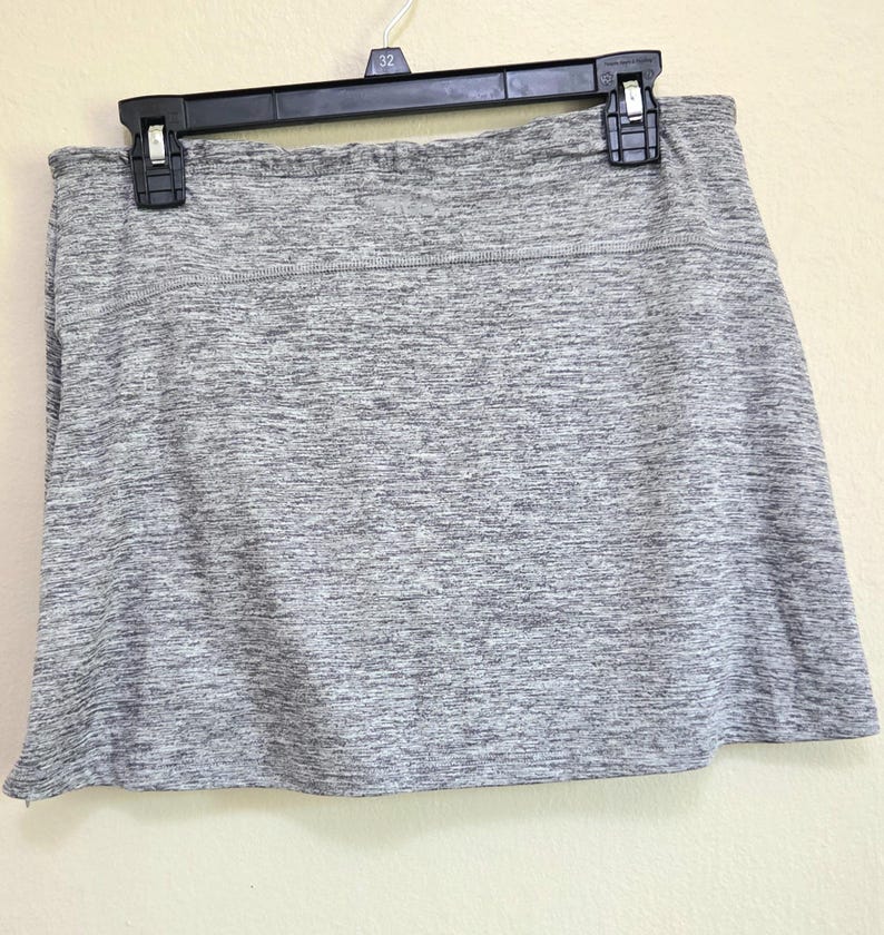 May include: A gray athletic skort hanging on a black hanger. The skort has a built-in shorts layer and a flared skirt. The fabric has a heathered appearance, with a mix of light and dark gray threads. The waistband is a solid gray.