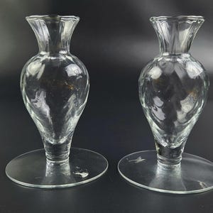 May include: Two clear glass vases with a bulbous body and flared rim. The vases have a textured surface and sit on a round, flat base. The background is a dark gray.