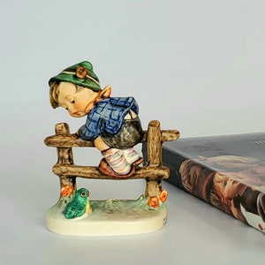 May include: A figurine of a boy wearing a green hat and blue and white clothing climbing a wooden fence. A small green frog figurine is at the base of the fence.