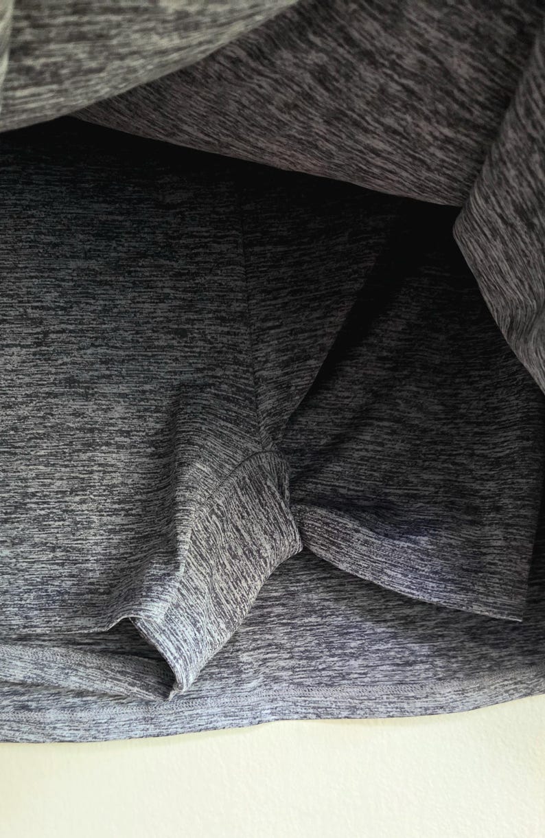 May include: Close-up of a gray athletic skort. The fabric has a textured, heathered appearance. The shorts portion is visible beneath the skirt, with seams and edges clearly defined. The garment appears to be made of a lightweight, breathable material.
