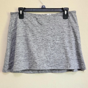 May include: Gray athletic skort hanging on a black hanger. The skort has a built-in shorts design and a wide waistband. The brand name "FILA" is visible on the waistband. The fabric has a heathered texture.