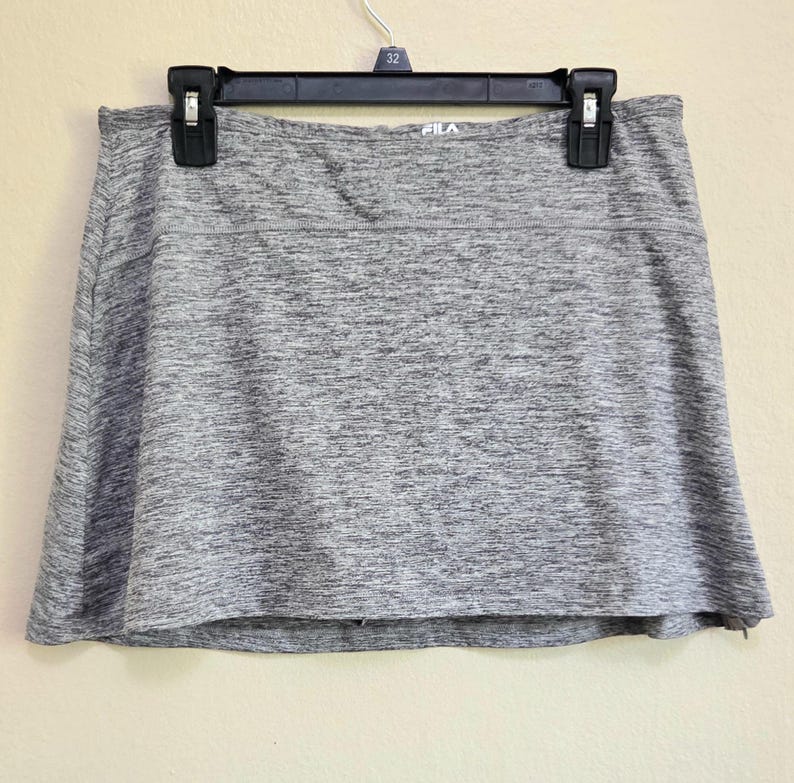 May include: Gray athletic skort hanging on a black hanger. The skort has a built-in shorts design and a wide waistband. The brand name "FILA" is visible on the waistband. The fabric has a heathered texture.