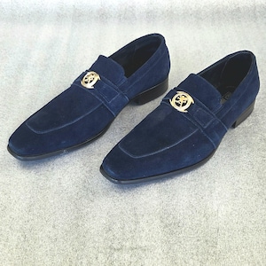Pode incluir: Um par de mocassins de camurça azul marinho com um emblema dourado na tira. Os sapatos têm bico quadrado e salto baixo. Os mocassins são exibidos em uma superfície cinza claro.
