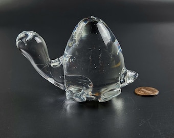 May include: A clear glass turtle ornament with a smooth, reflective surface. The turtle has a rounded shell, a head, and four flipper-like appendages. A penny is next to the turtle for size comparison.