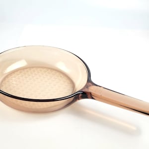 Puede incluir: Una sartén de vidrio marrón con un fondo texturizado y un mango largo.